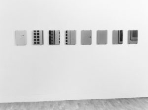 The photograph of ‘Imagination Dead Imagine’ is black and white. The artwork consists of 8 small rectangular mirrors, the type that wouldn’t look out of place hung above a bathroom sink, attached to the wall in a horizontal line. The wall surrounding the mirrors is completely blank. We can see reflections in some of the mirrors of what appear to be the doors into the room and the corner of another artwork. There is no reflection of the camera or photographer. There are no people in the photograph, either viewing the mirrors or reflected in them.