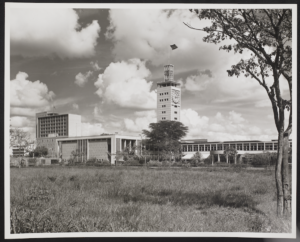 A photograph of the Nairobi parliament building, taken by Anthony Kersting. The photograph is black and white and shows the modernist clock down rising up from the low buildings. The photograph is catalogued as KER_PNT_G06606.