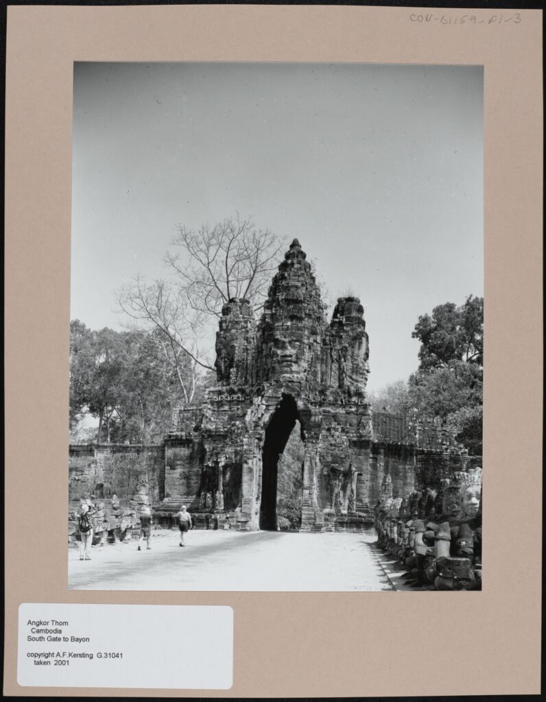 Black and white photograph mounted on card. The focus of the photograph is a large stone gate at the end of a wide, white path. At the centre of the gate is a carved stone face, underneath the face there is an archway flanked by two stone columns. Behind the gate there are many trees. There are several people walking towards the gate on the white path, which itself is flanked by rows of stone statues.