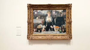 A photograph of the painting “A Bar at the Folies-Bergère”, by Édouard Manet. A young woman is looking out at the viewer, standing behind a bar and surrounded by bottles of alcohol, flowers, and a bowl of oranges. Behind her, the rest of the bar and its patrons are visible. The painting is framed by an ornate, carved wooden frame and hanging on a white wall.