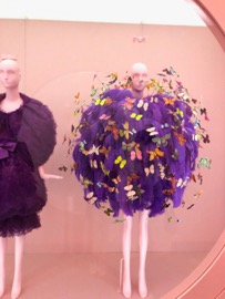 Two mannequins in exhibition, one wearing a purple, fluffy ensemble with butterflies. The other wearing a black dress. 