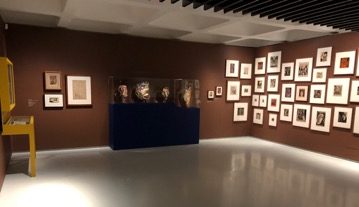 Exhibition at the Barbican, a brown wall with many framed woodcuts and a vitrine with four carved masks.