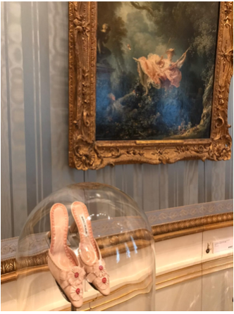 Light pink shoes with flowers in glass case beneath blue striped wall and painting of woman in pink dress