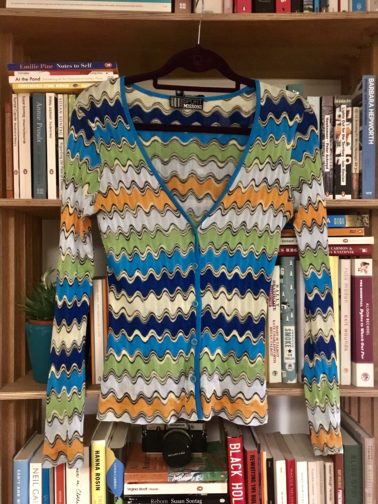 Colorful knit sweater hanging on wooden bookshelf filled with books. 