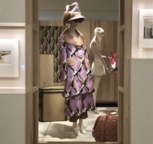 An image taken in an exhibition space, in which a mannequin dressed in elaborate, print ensemble by Louis Vuitton is positioned in the centre of the composition