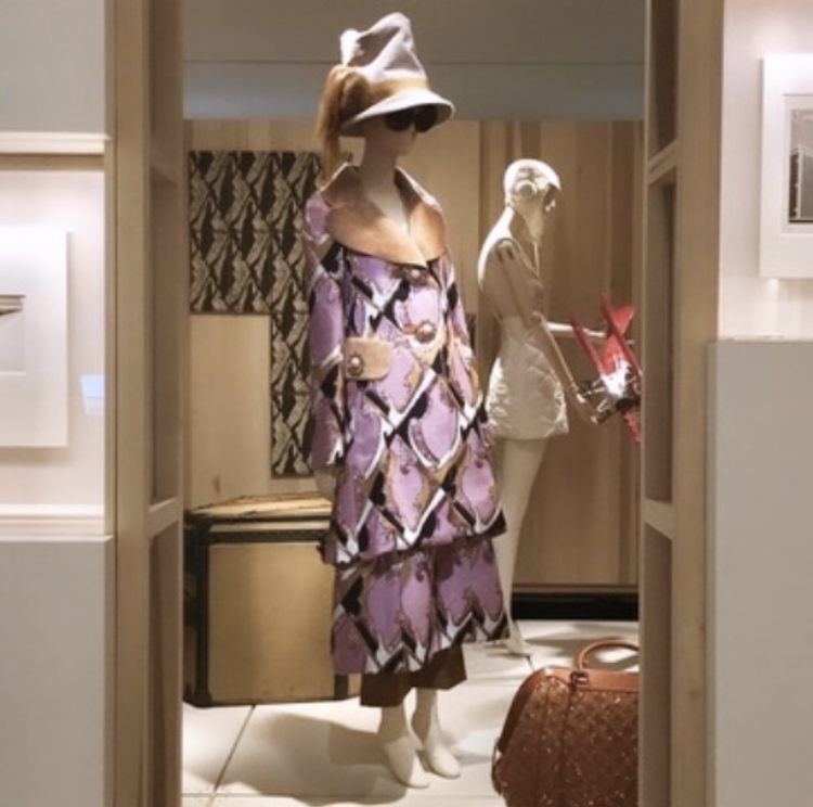 An image taken in an exhibition space, in which a mannequin dressed in elaborate, print ensemble by Louis Vuitton is positioned in the centre of the composition